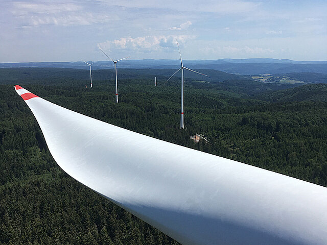 The rotor blade of a wind turbine is visible. The photo was taken from the turbine itself. In the background, more wind turbines are seen in the forest.