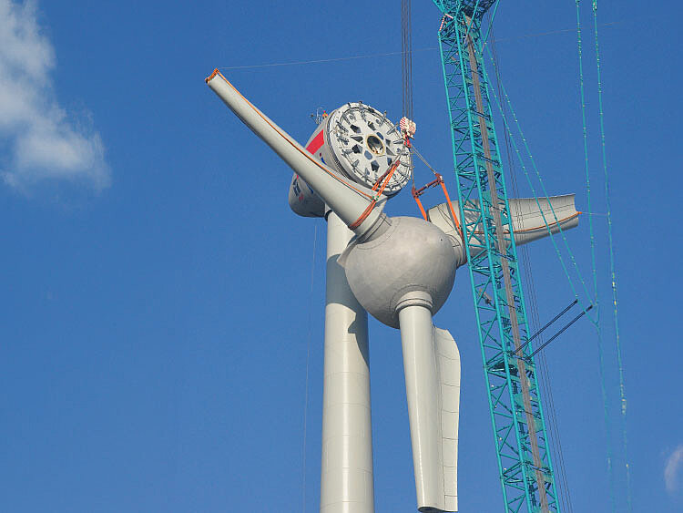 Using a construction crane, the nose of a wind turbine is being lifted to the nacelle.
