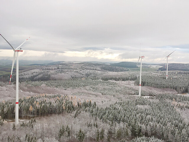The three turbines of the wind farm stand out against the winter landscape, blending in with the gray sky and snow-covered trees. The snow cover extends to the hills on the horizon.