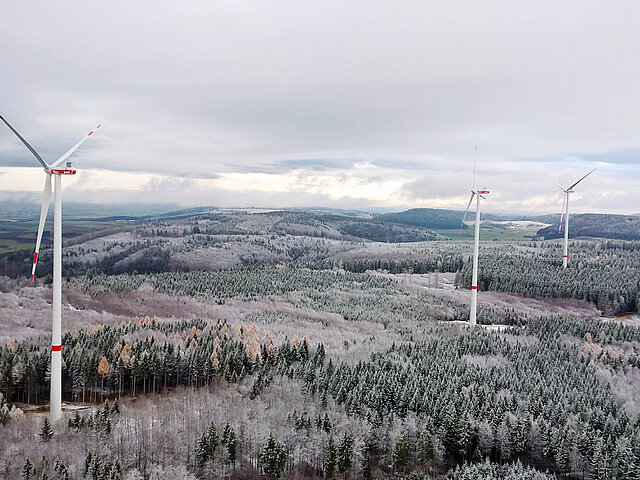 The three turbines of the wind farm stand out against the winter landscape, blending in with the gray sky and snow-covered trees. The snow cover extends to the hills on the horizon. 