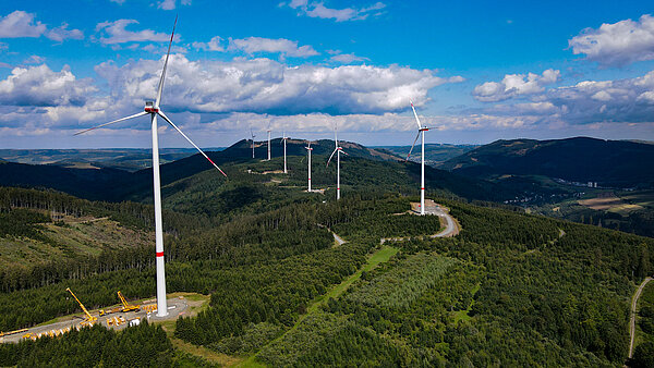 Seven wind turbines can be seen on a hilly, wooded area.