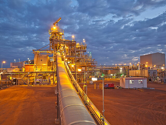 A conveyor belt of the mine in the twilight. Above the belt rises one of the production facilities. Everything appears illuminated by the many floodlights of the production facility.