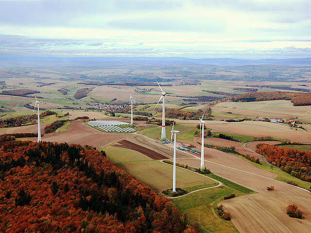 Between three smaller wind turbines, a significantly larger one towers. Another wind farm can be seen on the horizon.