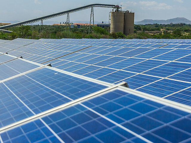 In the foreground, there are solar panels. In the background, there is a conveyor belt and other industrial facilities.
