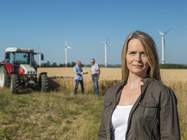 A JUWI employee is standing in a field. In the background, a farmer is standing next to his tractor, discussing with a colleague. Wind turbines are visible on the horizon.